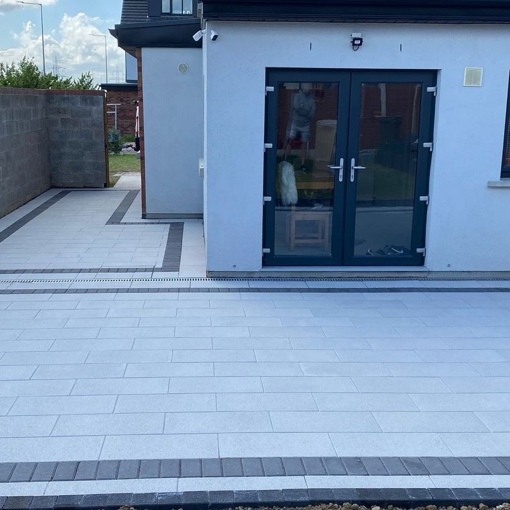 Paved patio with light gray and dark gray brick. A white building with double doors is on the right.