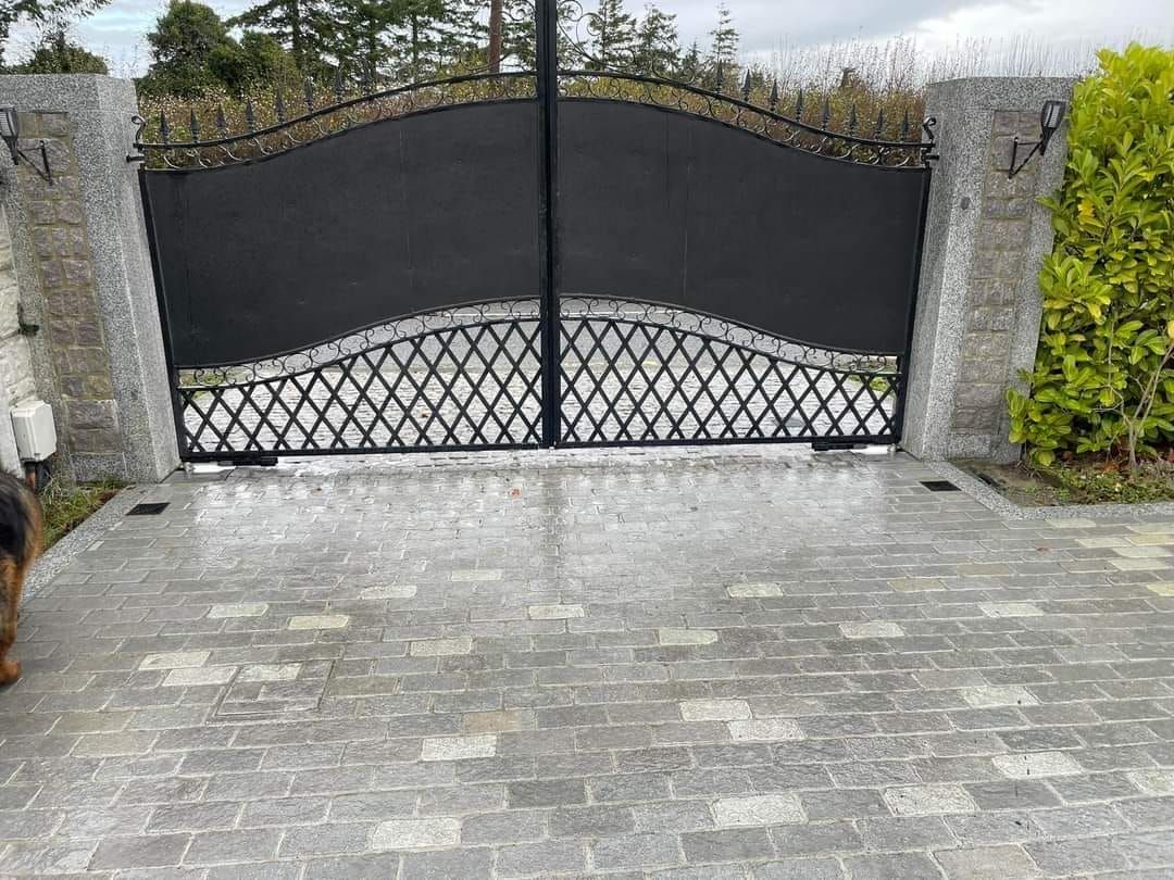 Black metal gate on gray brick driveway, flanked by stone columns and greenery.