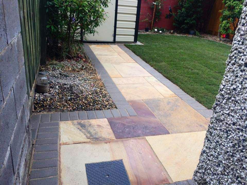Stone pathway lined with grass and a gravel bed next to a fence and a small shed.