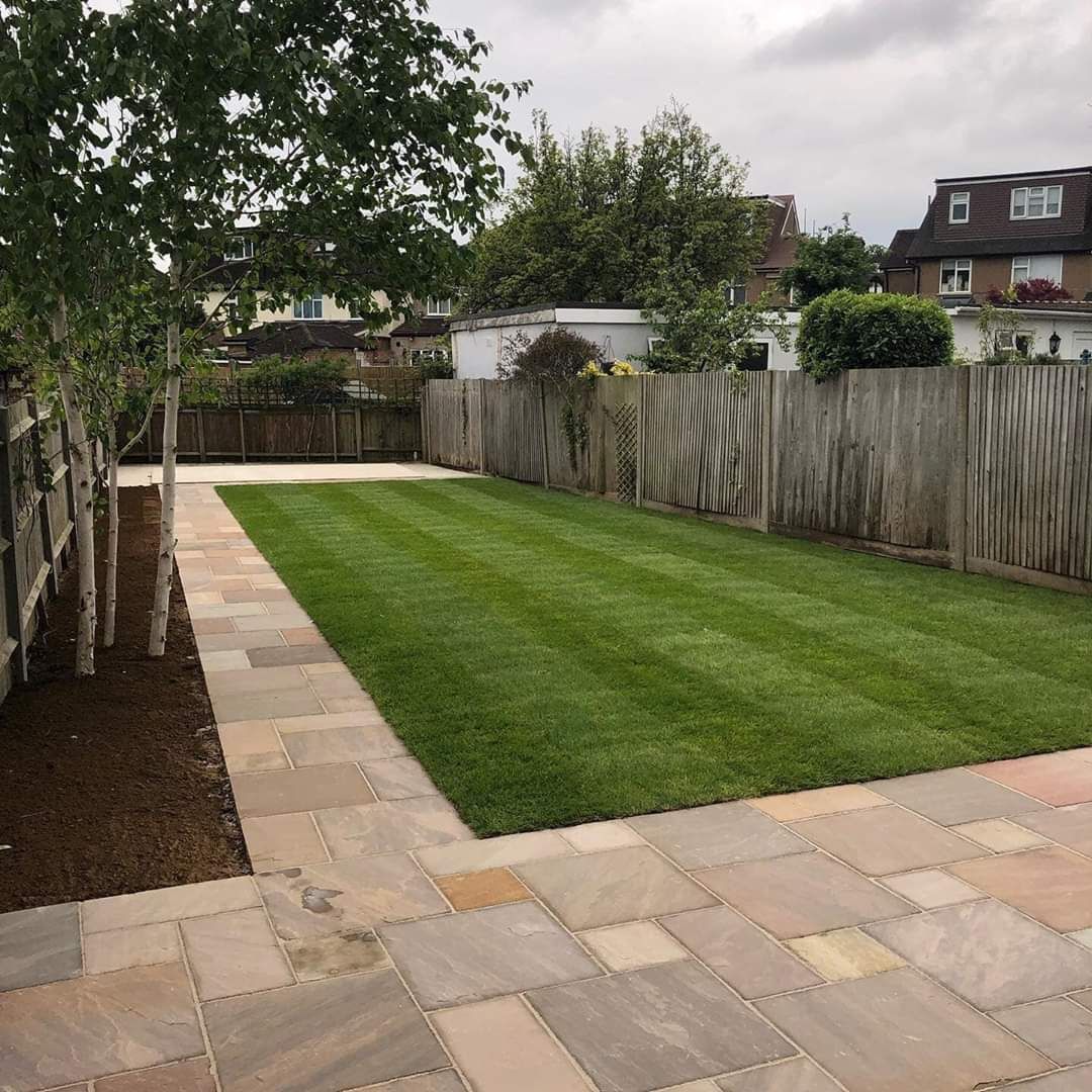 Backyard with a stone patio, manicured lawn, wooden fence, and trees.