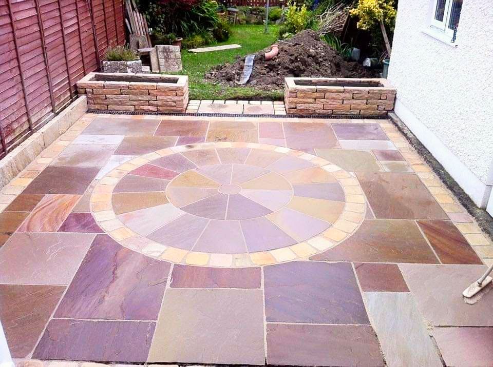 Stone patio with a circular design in a garden, with flower boxes and a fence.