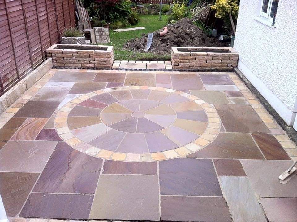 Patio with circular stone design and rectangular paving, bordered by planters and a fence.