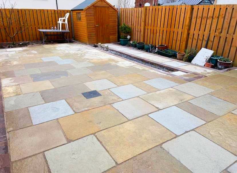 Stone patio with wooden shed and fence in a backyard. Various tan and gray flagstones.