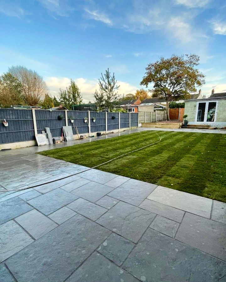 Stone patio leads to a freshly mowed green lawn with a gray fence and cloudy blue sky.