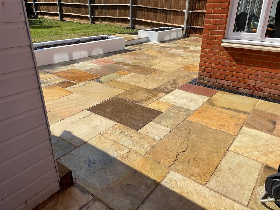 Patio with sandstone tiles in a garden, next to a brick wall, fence, and a white planter.