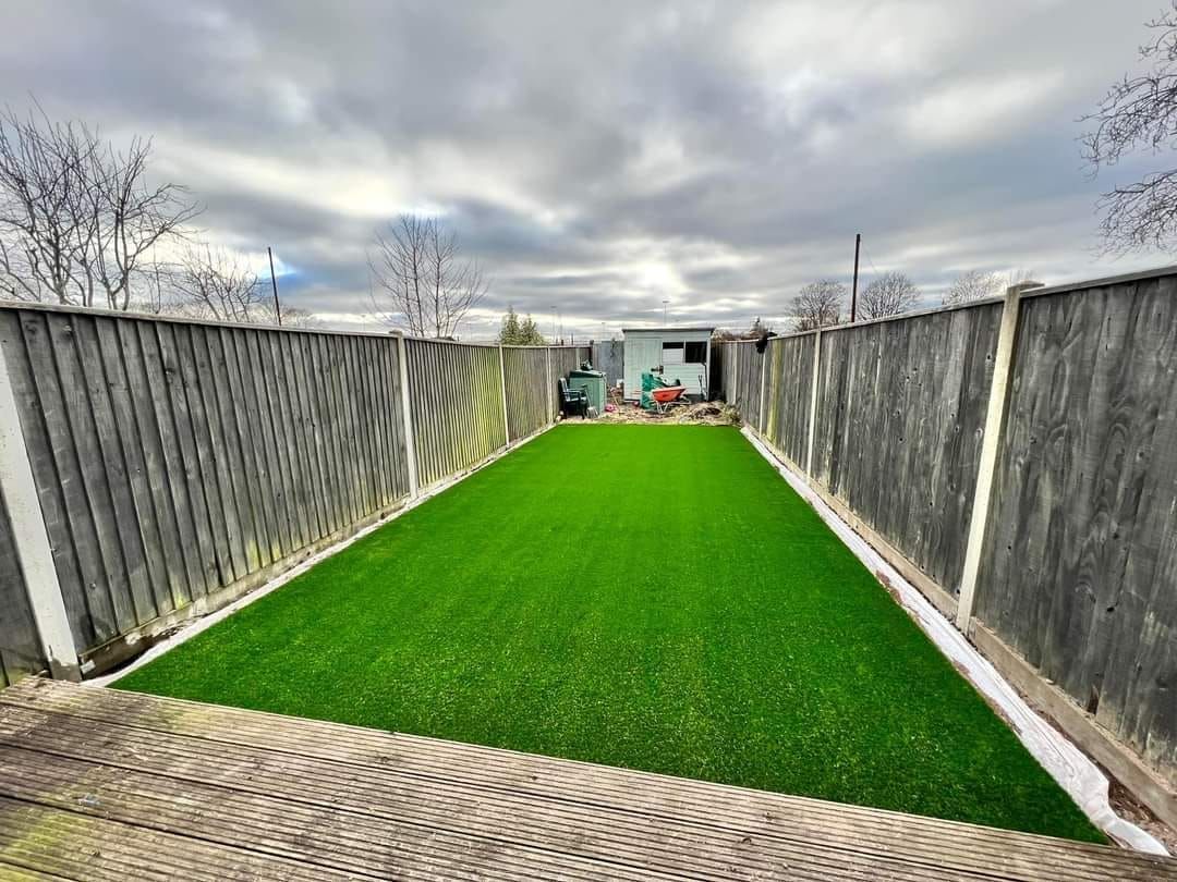 Rectangular backyard with green artificial turf, bordered by gray fences and a cloudy sky.