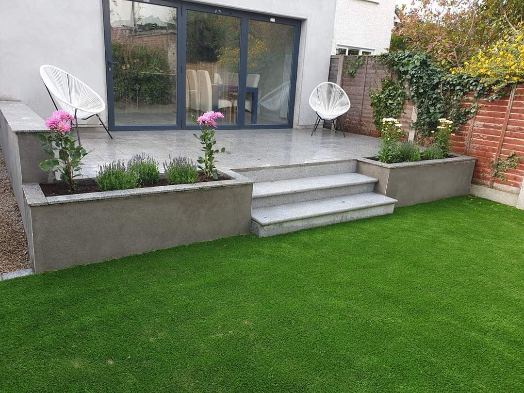 Backyard patio with steps, planters, and green lawn. Glass doors, two white chairs, and pink flowers.