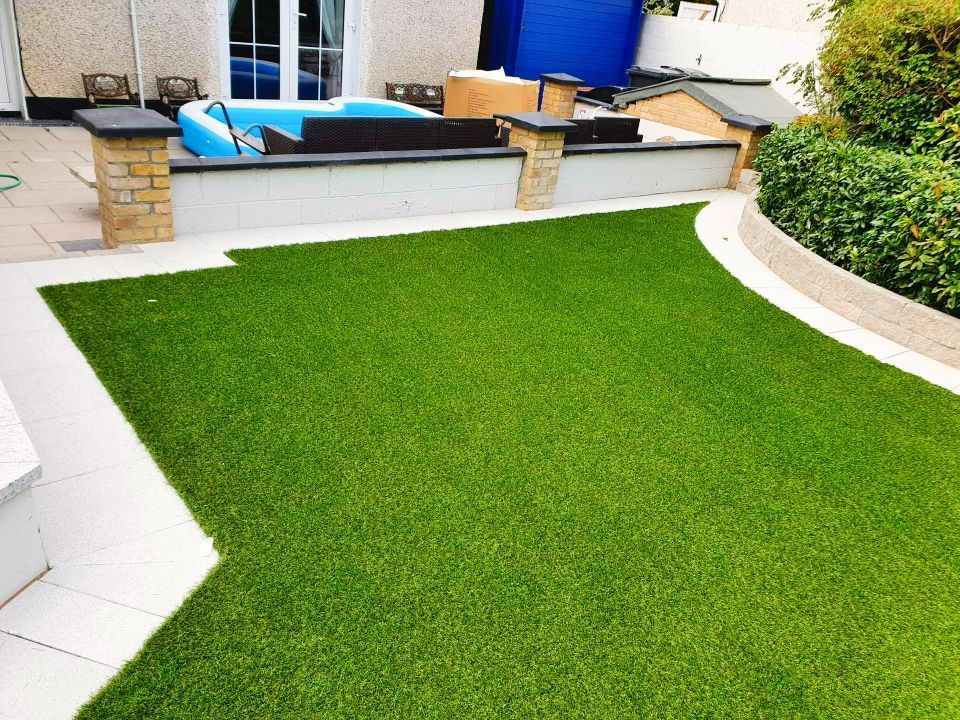 Green artificial lawn in a backyard, bordered by paving stones and a low wall.