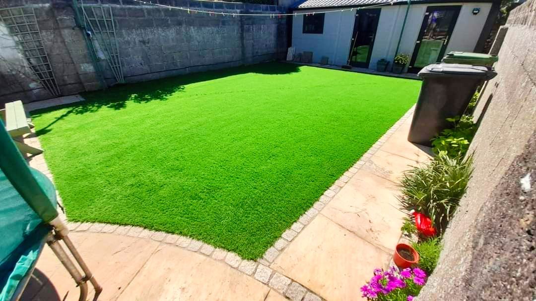 Green artificial lawn in a backyard, surrounded by stone and a small building.