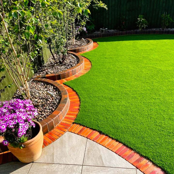 Lush green lawn bordered by brick and flowerbeds, with a potted purple flower in the corner.