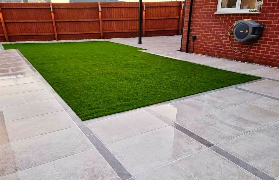 A rectangular backyard with artificial turf surrounded by light-colored paving stones.