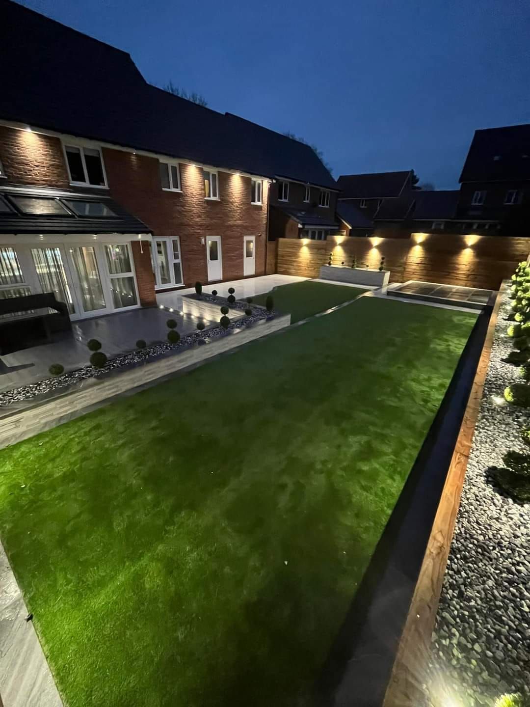 Backyard at dusk, with lit lawn, patio, bordering walls, and house with lights.