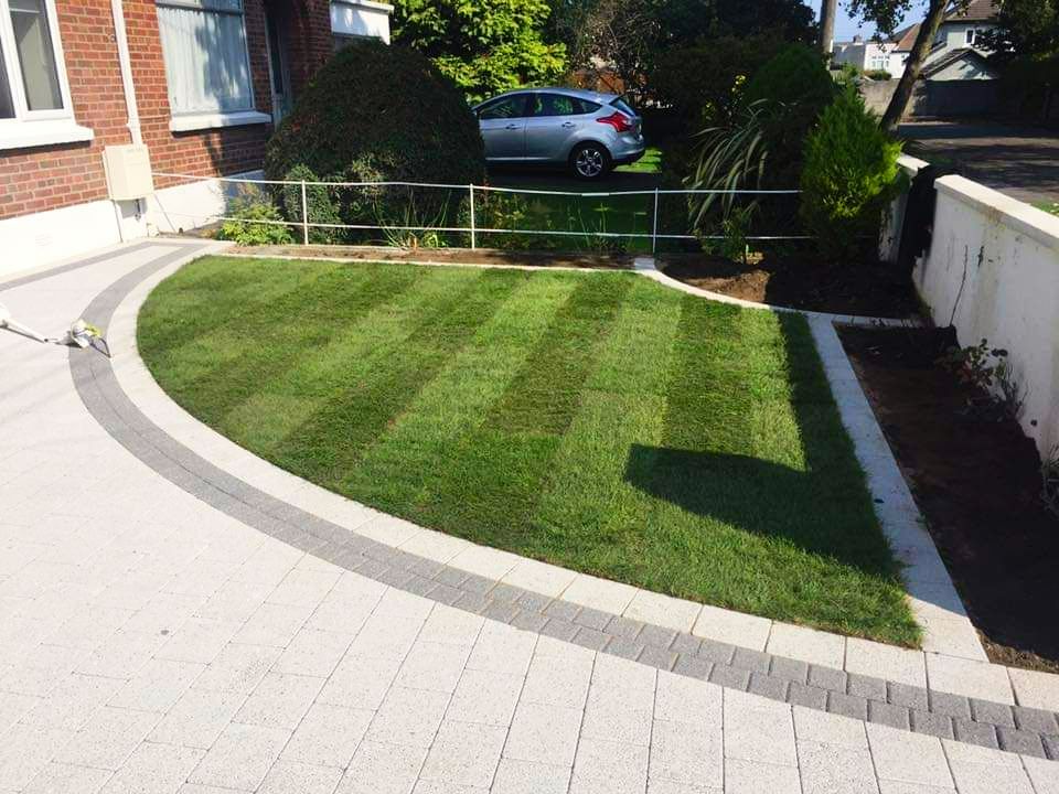 Lawn with stripes next to a curved driveway with paving stones. A car is parked in the background.