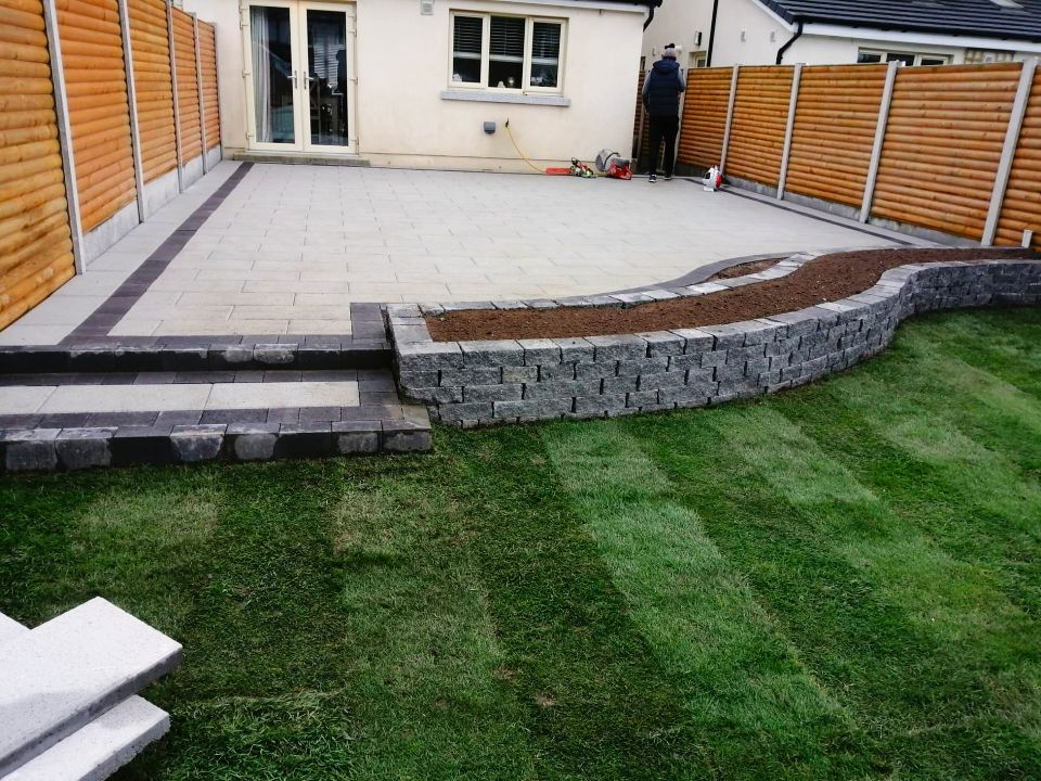 Patio with stone wall and steps, adjacent to a lawn and wooden fence.