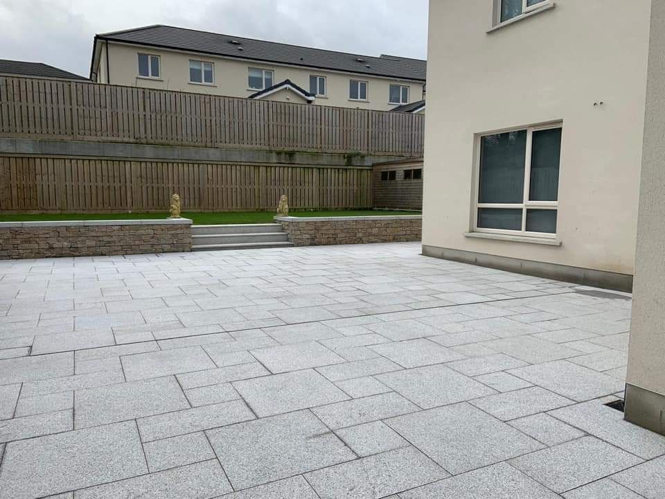 Gray paved patio in a yard, leading to a lawn and wooden fence. A cream building is on the right.
