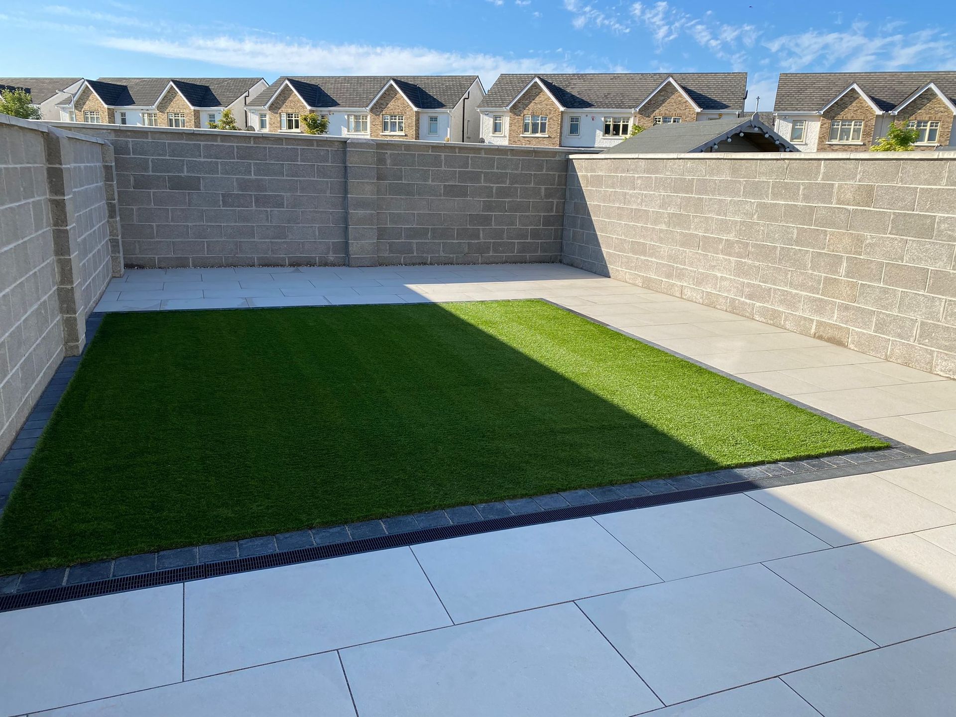 A small backyard with a strip of green grass and paved patio bordered by block walls, with houses in the background.