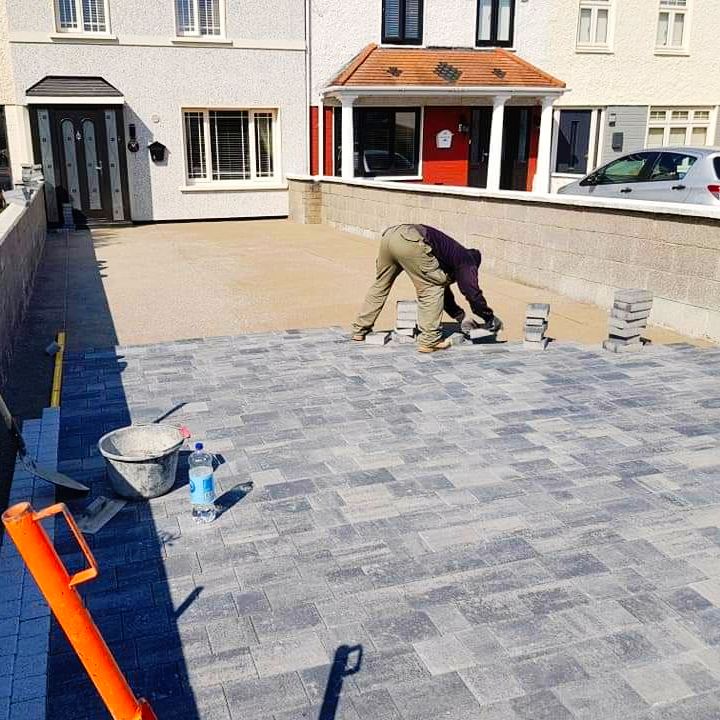 A person laying gray paving stones in a driveway. Construction site with tools and blocks visible.