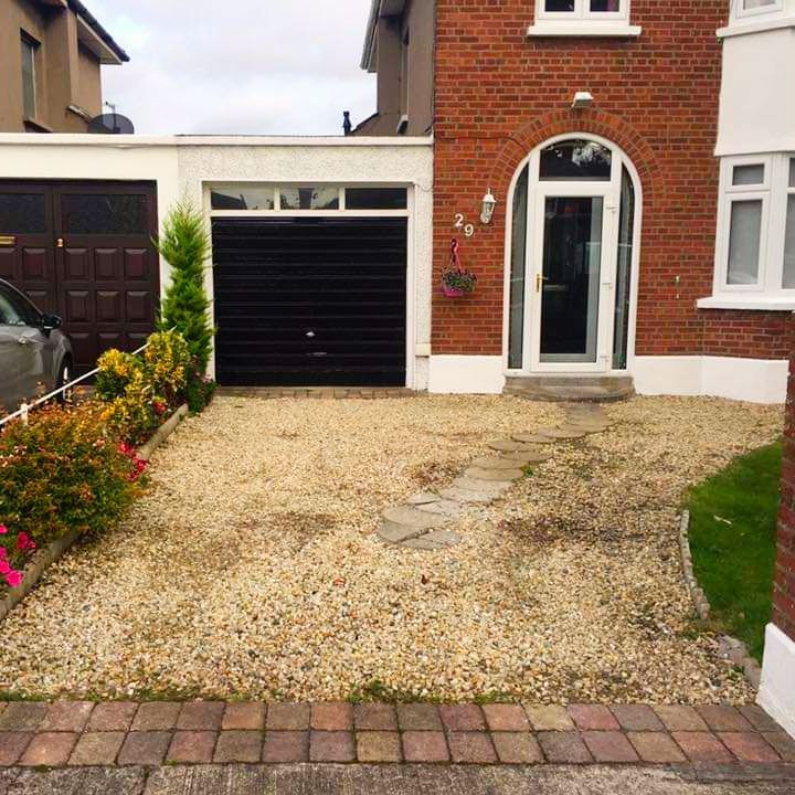 Driveway with gravel, plants, garage, and a brick house with a front door.