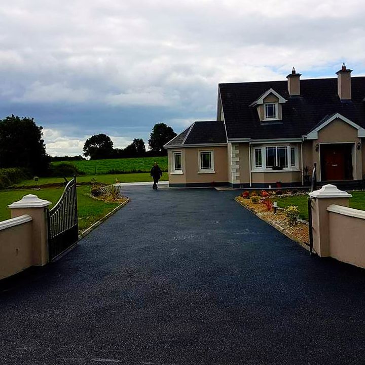 Black asphalt driveway leading to a beige house, person walking, green field in background.