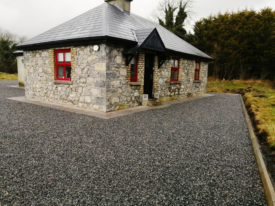 Stone cottage with dark roof and red window frames. Gravel driveway.