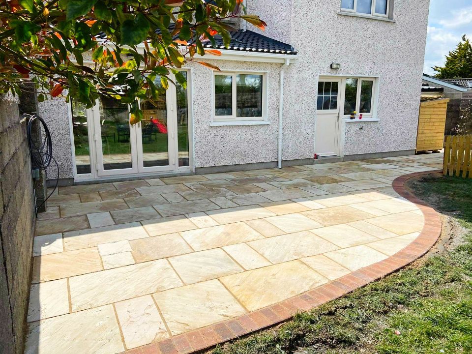 Paved patio with tan rectangular stones, bordered by red brick and grass, next to a cream house.
