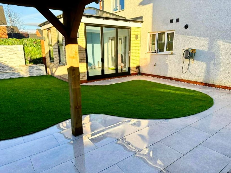 Backyard with green turf, patio, and a pergola. Sunlight, with house and sliding glass doors visible.