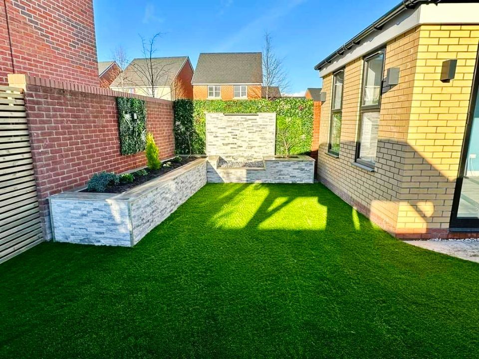 Backyard with brick walls, faux grass, a water feature, and greenery against a bright blue sky.