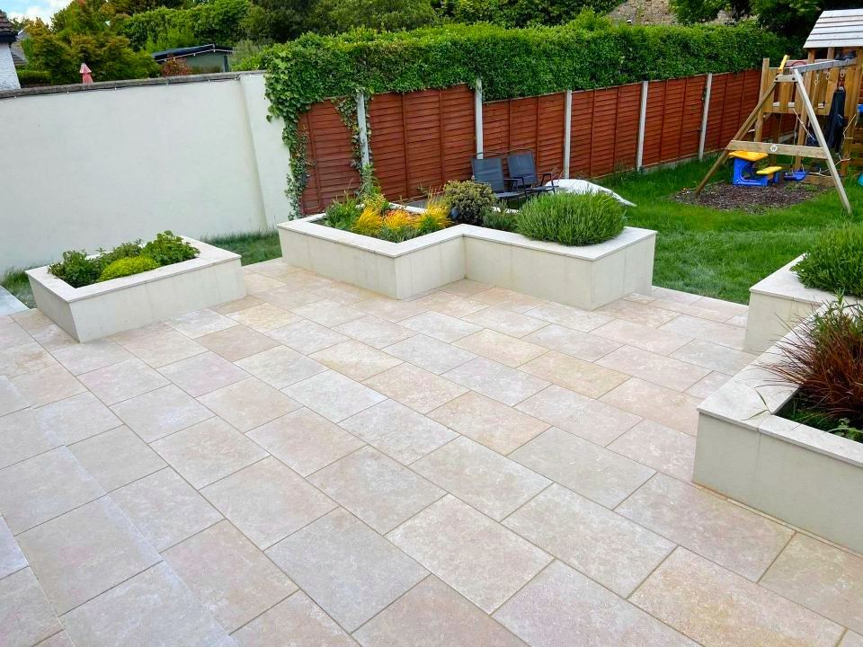 Patio with rectangular tan paving stones and built-in planters with greenery. Garden in background.
