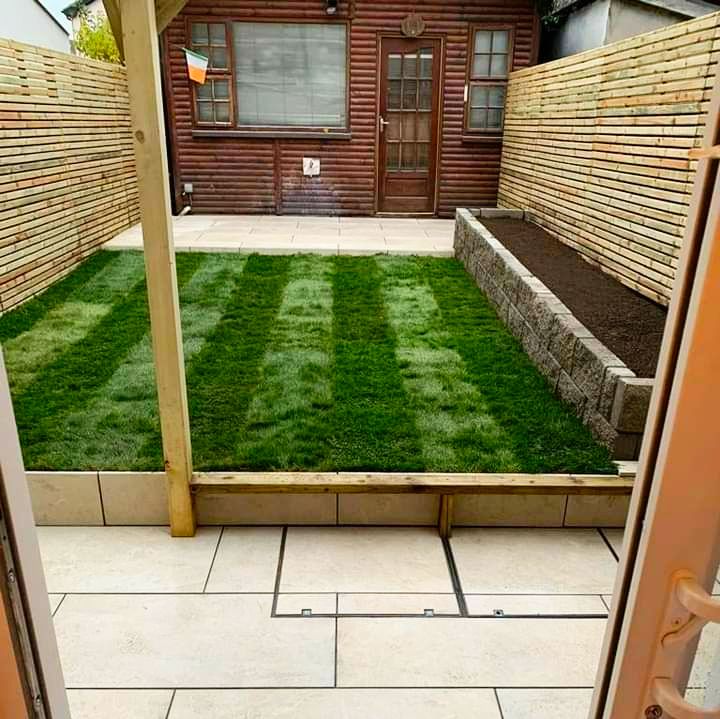 Small backyard with striped green lawn, stone patio, wooden shed, and slatted fence.