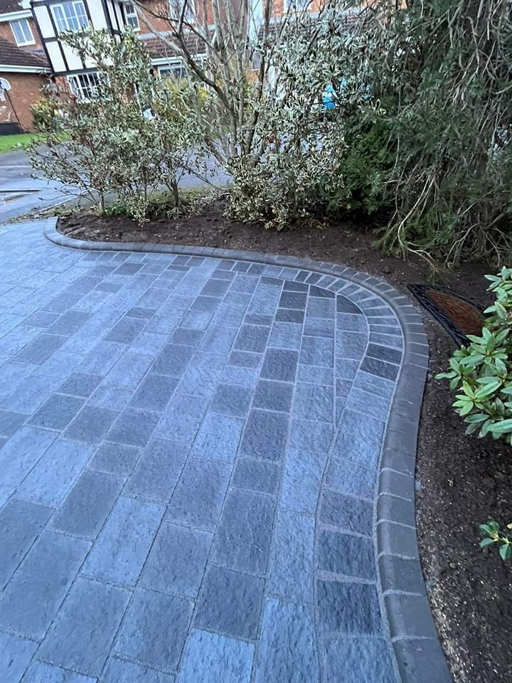 Dark gray brick driveway with curved edge, bordering soil and shrubs.