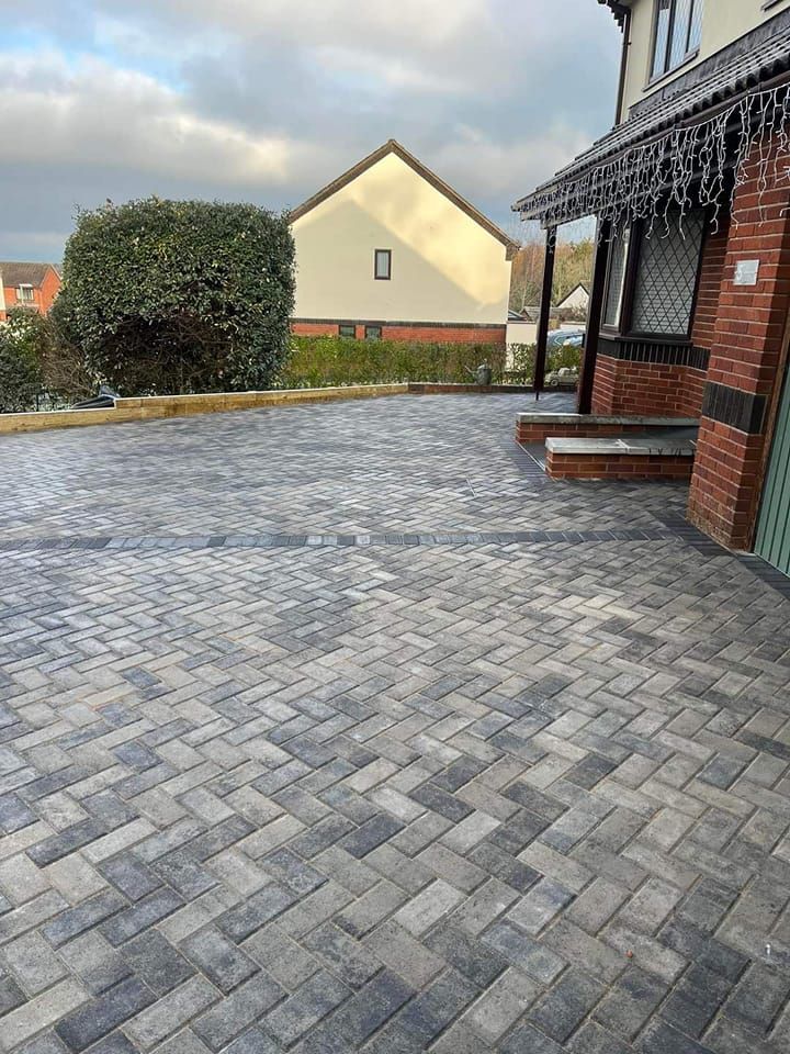 Herringbone patterned brick driveway in front of a house, leading towards a hedge and a building with a peaked roof.