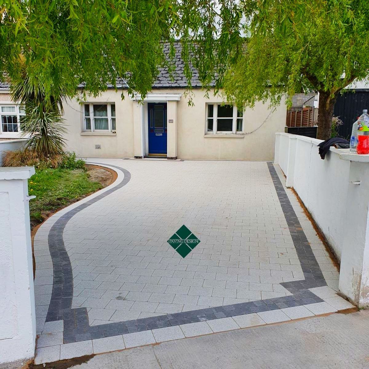 White cottage with a patterned driveway. Blue door, green trees, gray and white paving.
