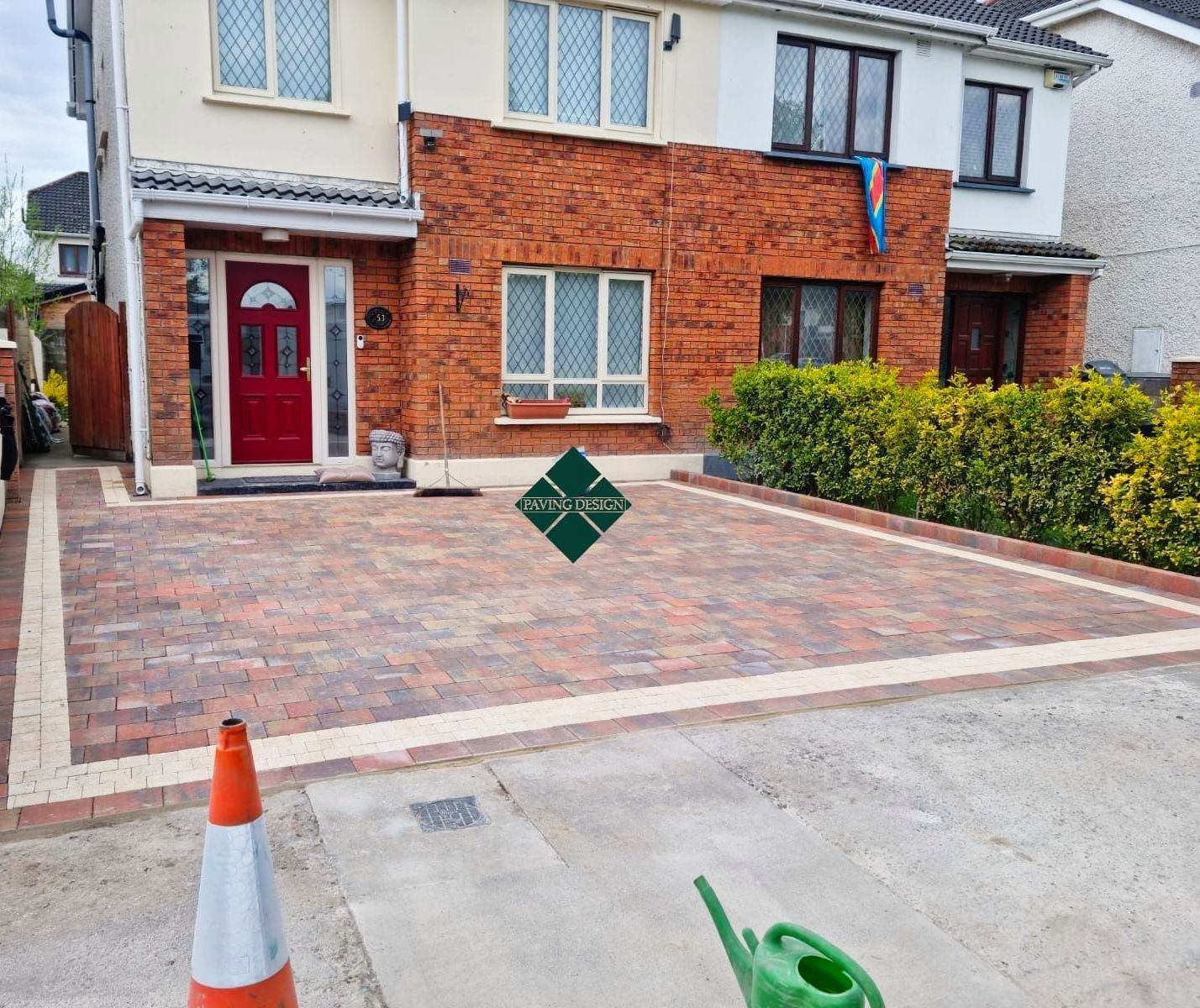 Brick driveway in front of a house, orange traffic cone, green watering can.