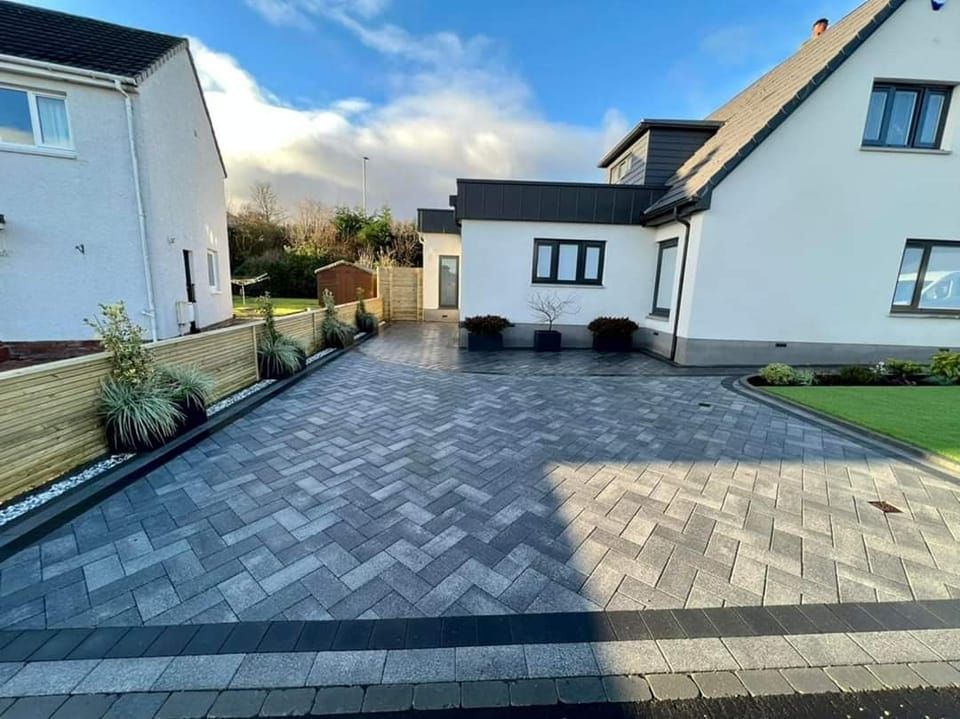Driveway with herringbone pattern pavers, bordered by dark pavers and green lawn, with white house.