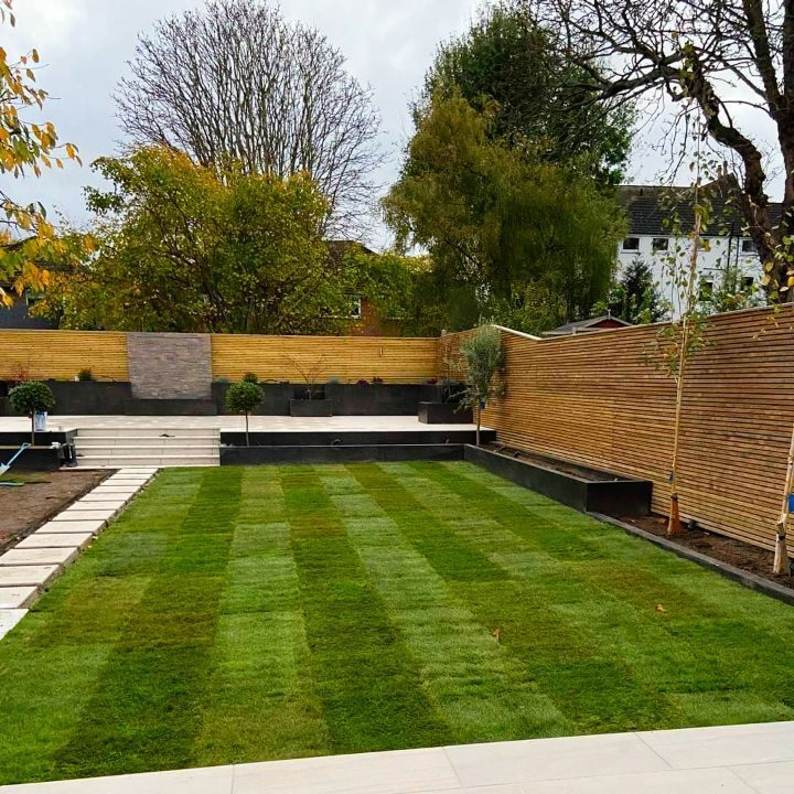 Green lawn with striped mowing pattern, tiered patio, wooden fence, trees, and cloudy sky.