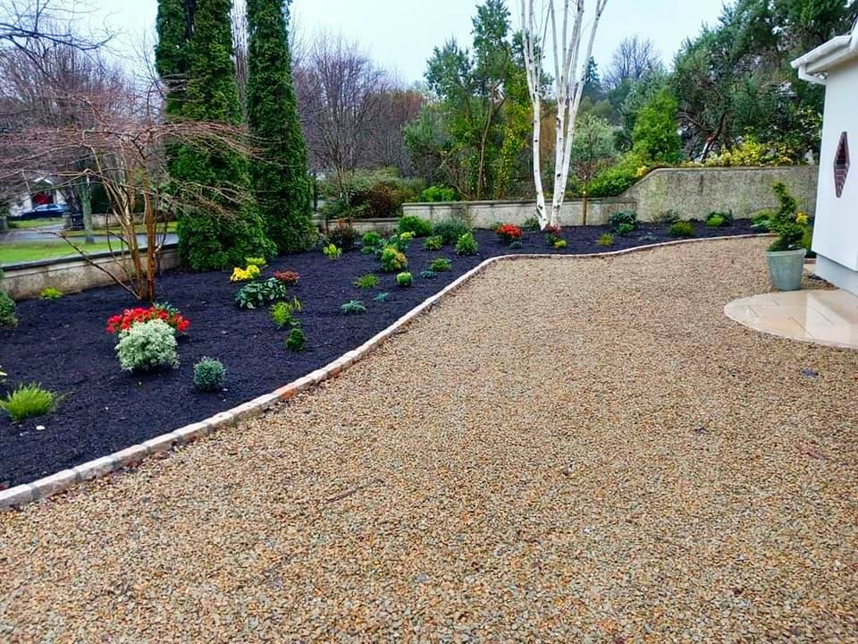 Gravel driveway borders a landscaped garden with dark mulch and various plants, next to a white house.
