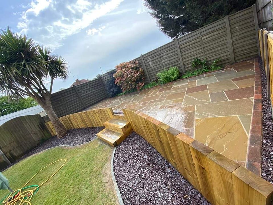 Patio with stone pavers, wooden retaining walls, steps, and a grassy area with a tree and fence.