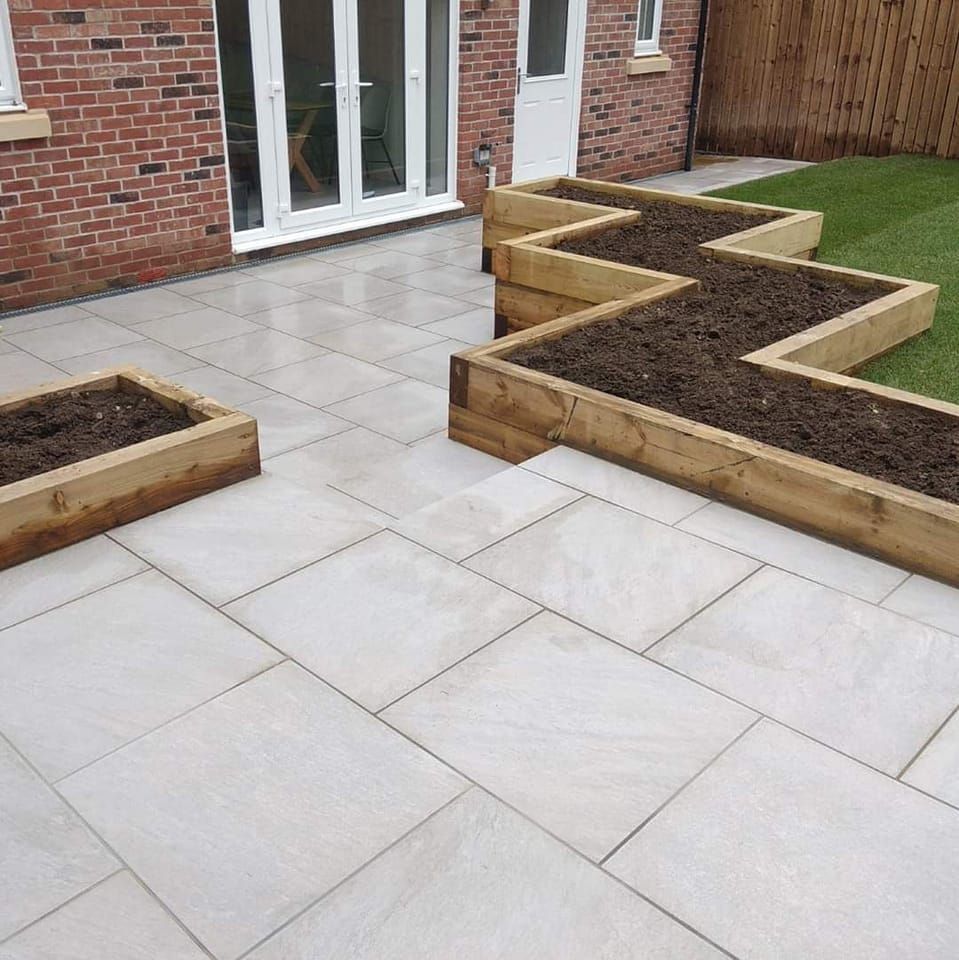 Patio with light gray paving stones, wooden zig-zag planter boxes filled with soil, and a green lawn.