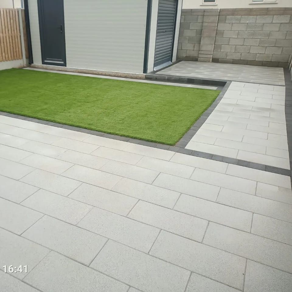 A backyard with artificial green grass, light gray pavers, and a white shed with a dark gray door.