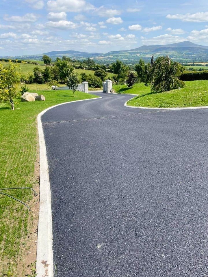 A paved driveway curves through green lawns, leading to pillars and a scenic hillside view.