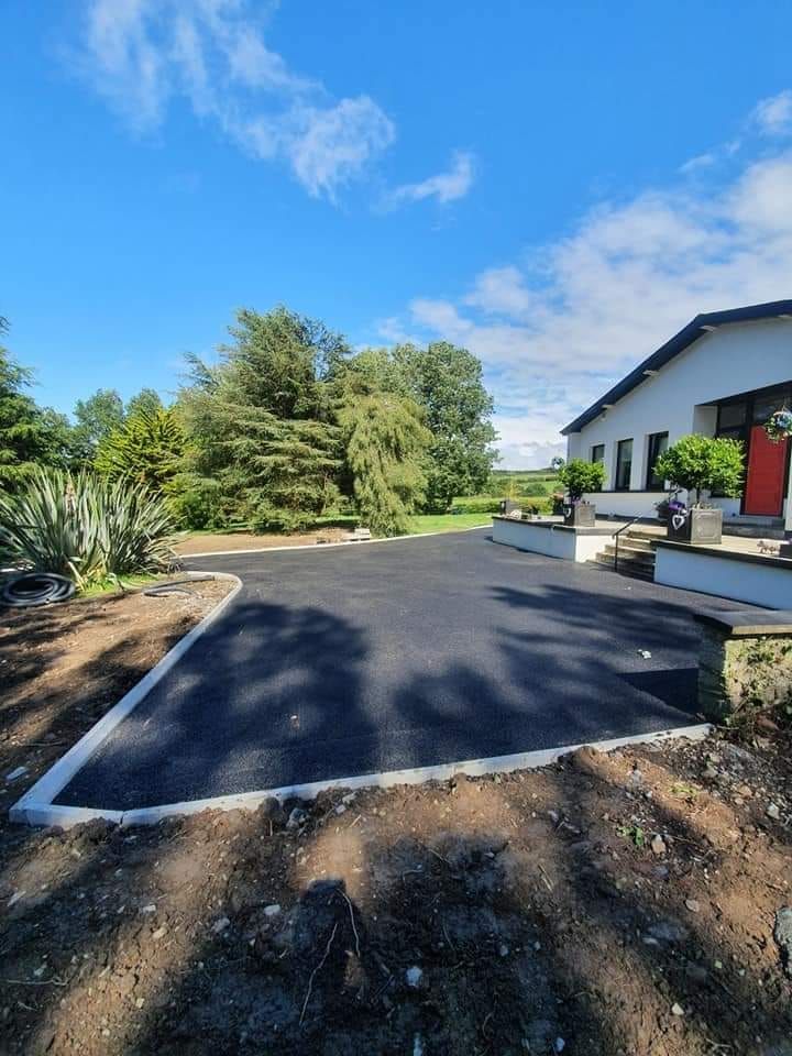 Newly paved asphalt driveway with concrete edging in front of a white house on a sunny day.