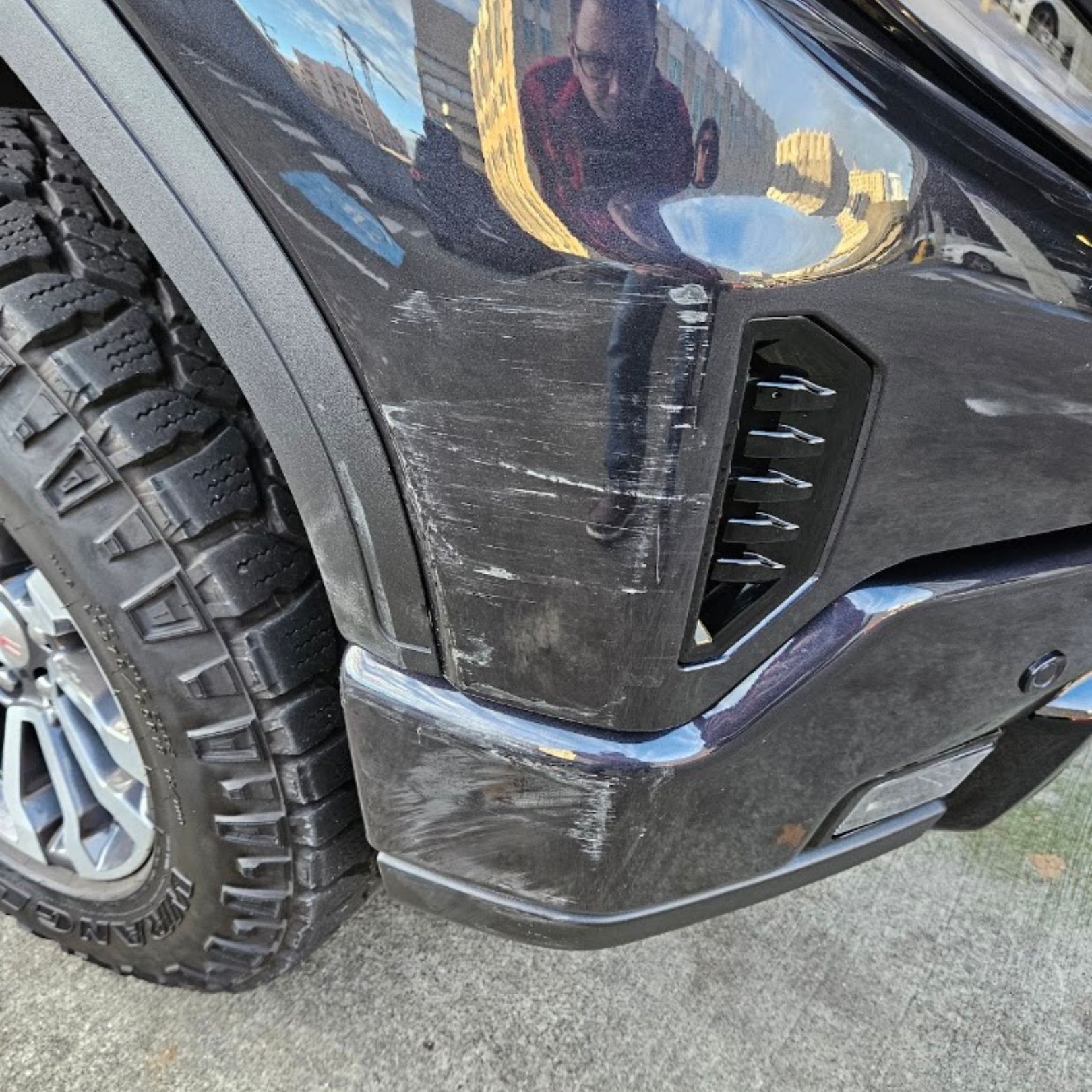 Close-up of a black SUV’s front wheel and bumper, showing scratches and scuffs on the side panel.