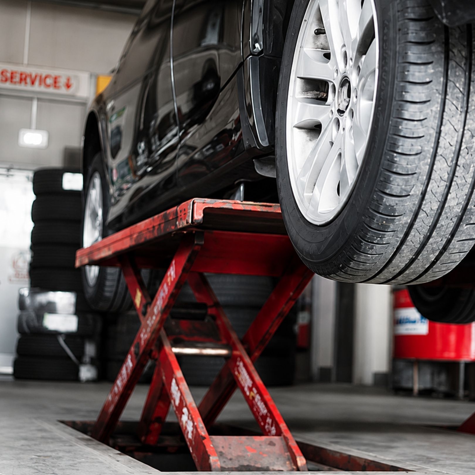 Black car raised on a red scissor lift in an auto repair shop, with a tire in the foreground