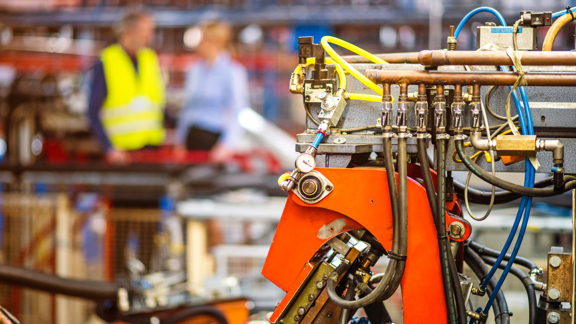 Industrial machinery in a factory, with a blurred worker in a yellow safety vest in the background.