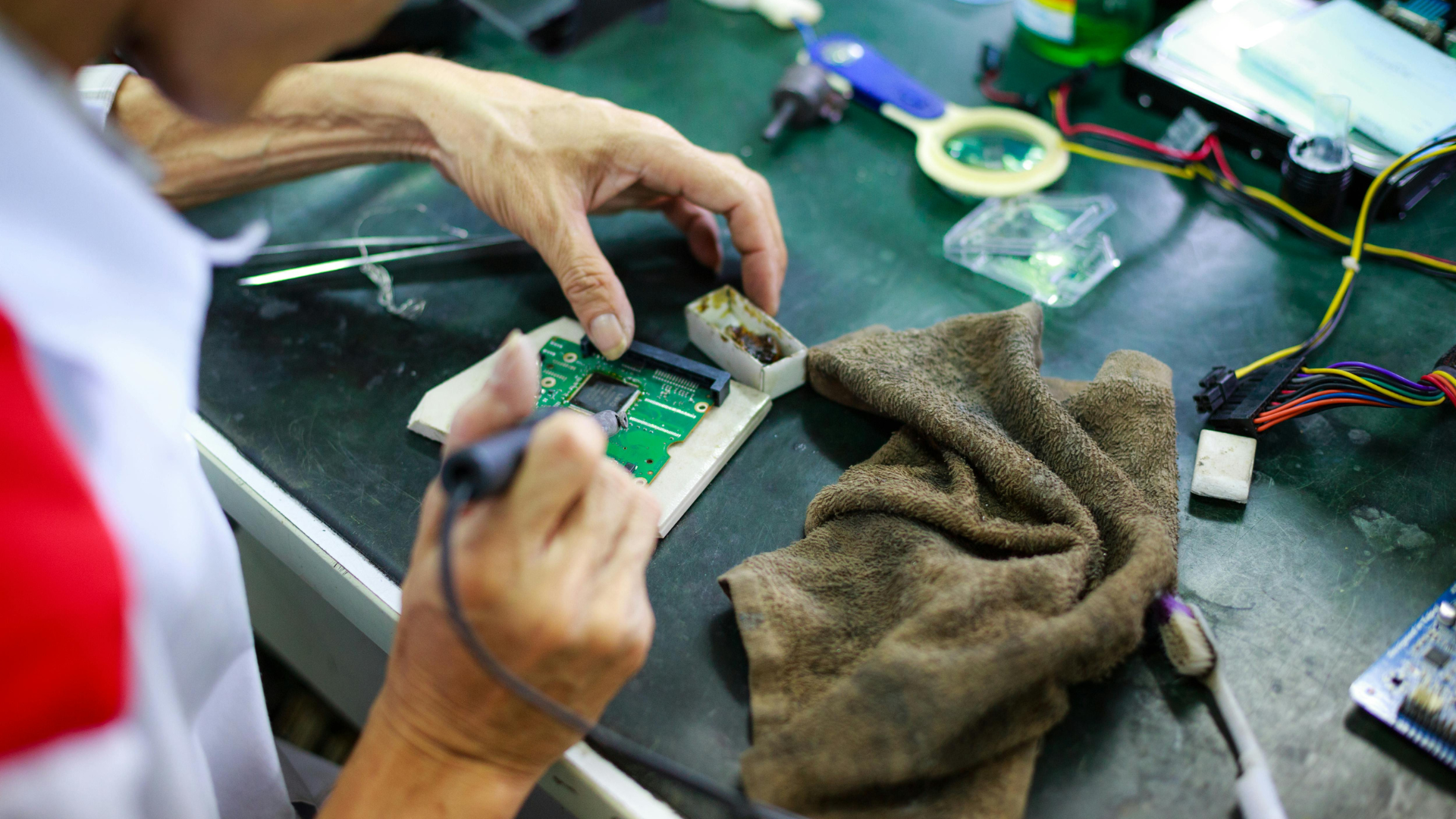 Hands using a tool to repair a green circuit board on a workbench with wires and cloth