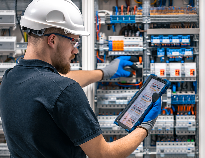 Technician in hard hat inspecting an electrical control panel with a tablet and blue gloves