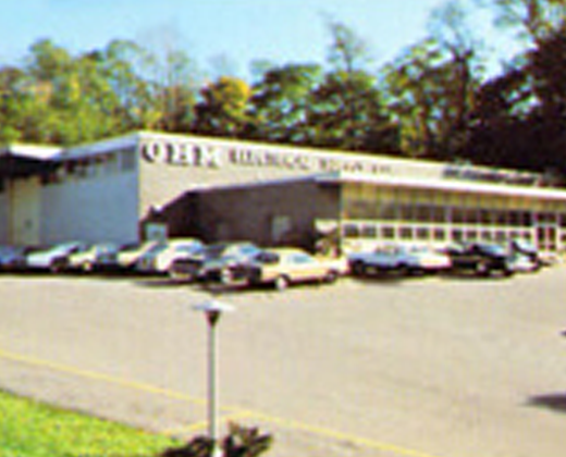 Parking lot outside a low white office building with several parked cars and trees behind it