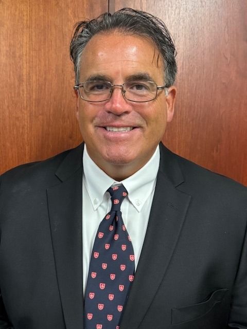 Man in a dark suit and patterned tie smiling against a wood-paneled background