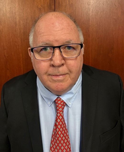 Man in a black suit, blue shirt, and red tie, wearing glasses, stands against a wooden background.