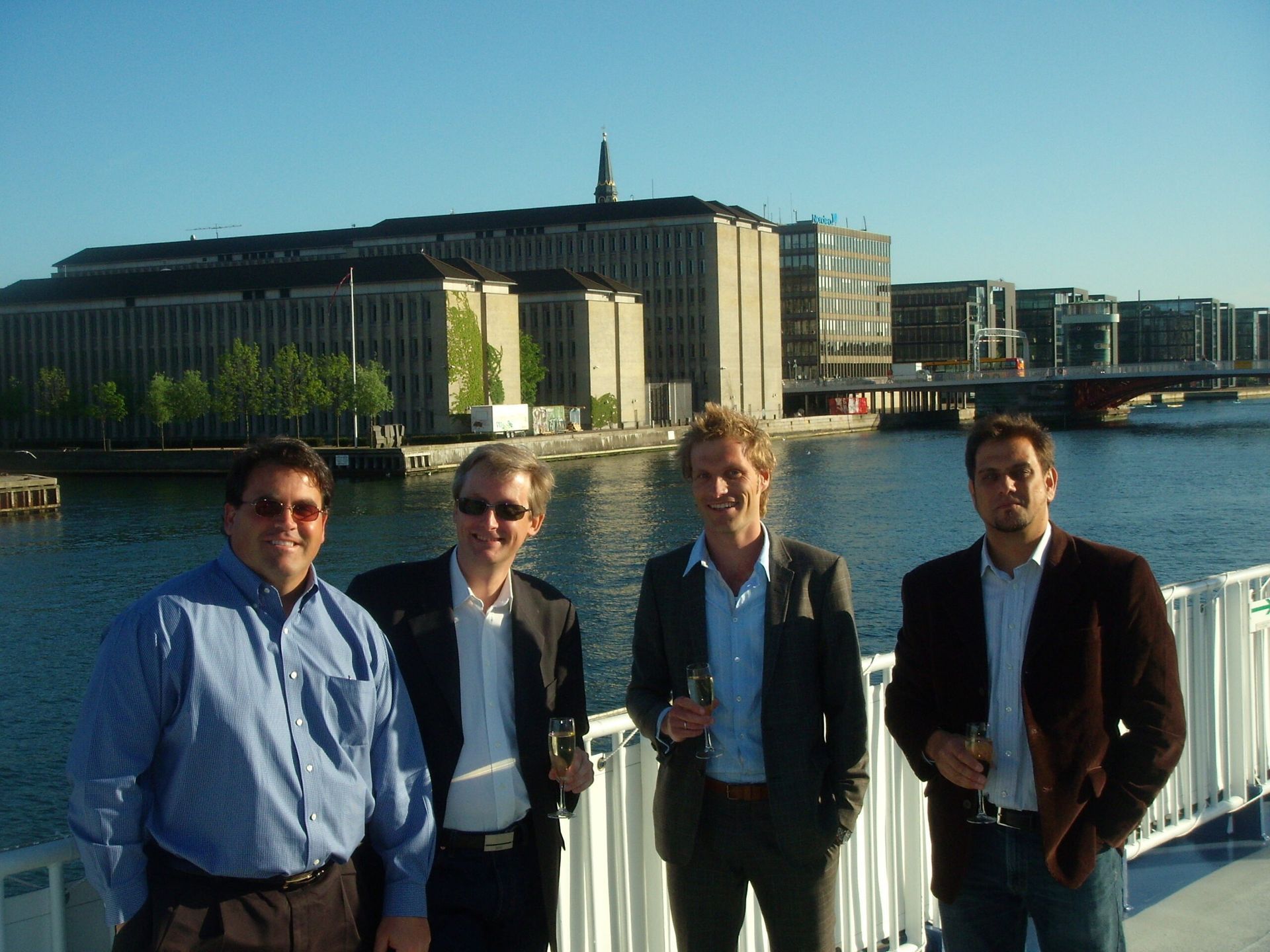 Four people standing by a waterfront with city buildings and a bridge in the background